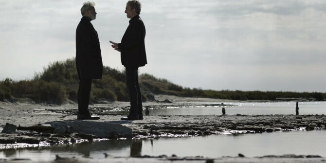 Deux hommes en manteau sombre discutant sur un rivage rocheux sous un ciel gris – série Ici tout commence